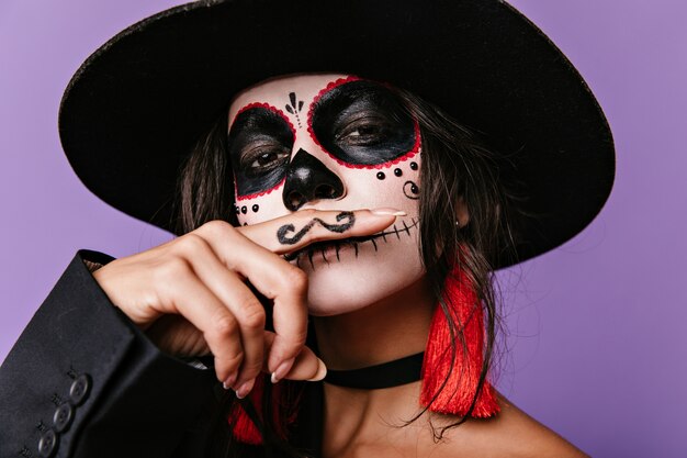Foto de una chica brillante con sombrero de ala ancha que representa a un hombre mexicano con bigote. Señora morena posando en la pared lila.