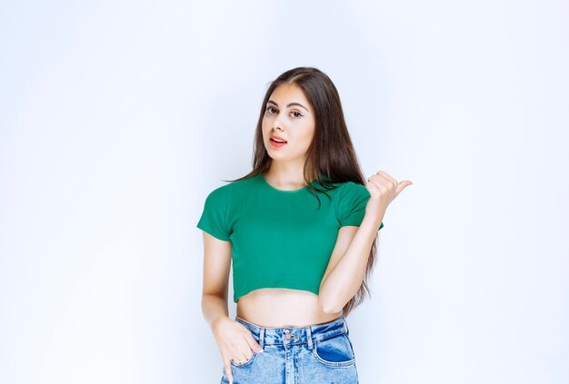 Foto de una chica guapa modelo de pie y apuntando a un lado contra la pared blanca.