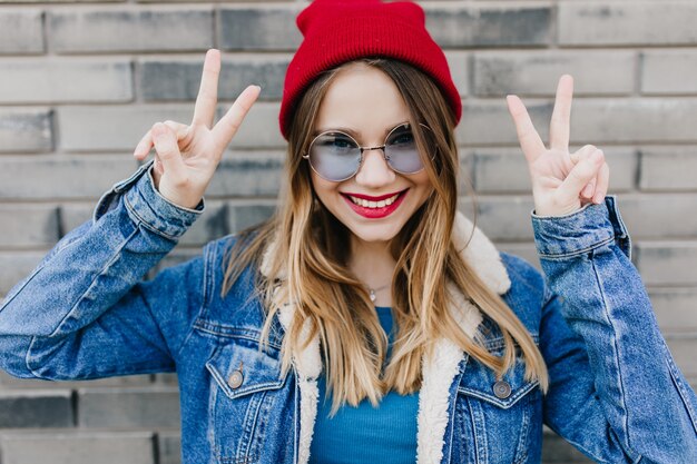 Foto de una chica increíble en vasos redondos expresando felicidad en la pared de ladrillo. Tiro al aire libre de mujer blanca de ensueño con sombrero rojo y chaqueta vaquera riendo
