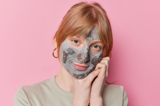 Foto gratuita la foto de una chica pelirroja seria se apoya en las manos aplica una máscara de arcilla de belleza en la cara para el tratamiento de la piel tiene una mirada tierna a las poses de la cámara contra un fondo rosa peeling de cuidado facial y concepto de bienestar