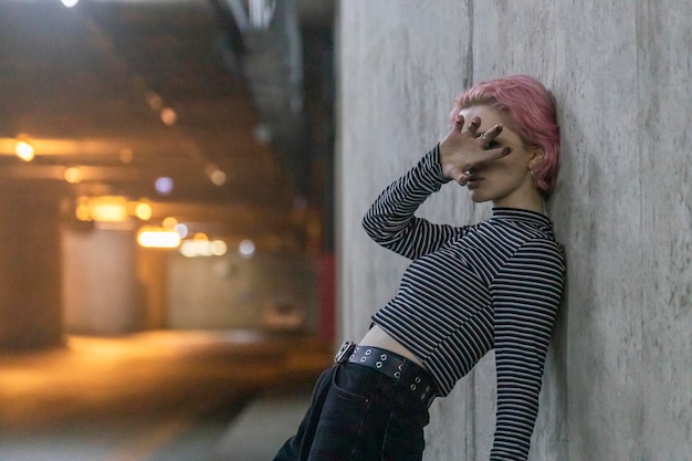 Foto de una chica de pelo rosa apoyada en la pared de la calle y llevándose la mano a la cara