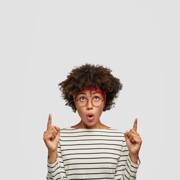 Foto de chica de piel oscura estupefacta con corte de pelo afro, mantiene la boca redondeada