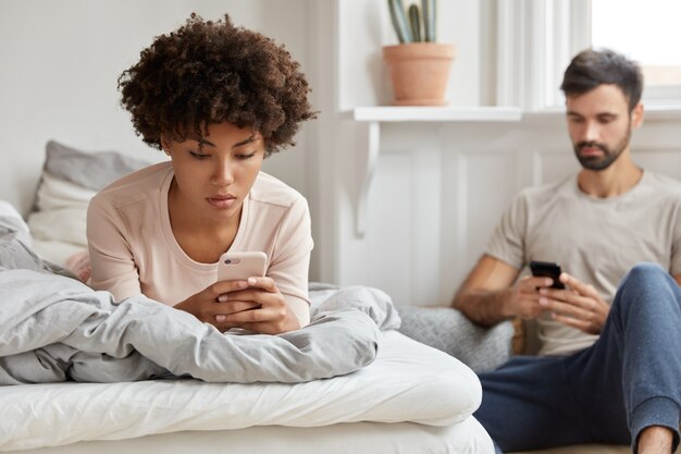 La foto de una chica relajada y despreocupada en pijama verifica la notificación en el teléfono móvil, descansa en la cama, su novio barbudo se sienta en la pared, envía un mensaje de texto a un amigo, está conectado a internet inalámbrico