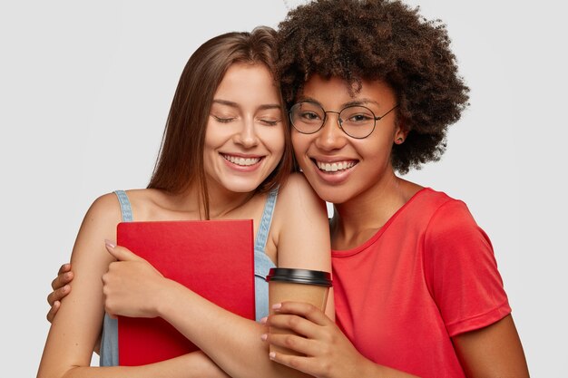 Foto de chicas positivas amistosas de raza mixta abrazan mientras pasan tiempo juntos estudiando