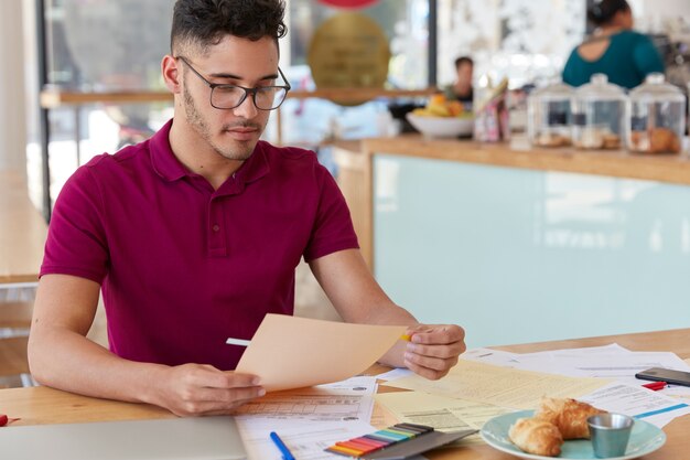 Foto de chico sin afeitar examina el papeleo, usa pegatinas, vestido con una camiseta informal y gafas. Blogger masculino creativo trabaja con documentación, tiene un día de trabajo duro, desarrolla una nueva estrategia.