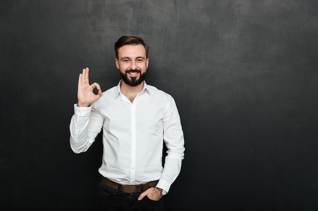 Foto de un chico sin afeitar en la oficina sonriendo y gesticulando con un signo OK expresando que todo está bien, aislado sobre grafito