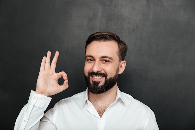 Foto de chico barbudo sonriendo y gesticulando con un signo OK expresando una buena elección, aislado sobre grafito