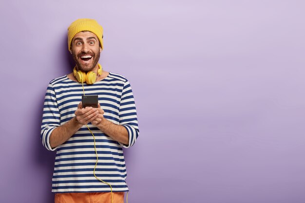 Foto de un chico inconformista sin afeitar, feliz y energizado, sostiene un teléfono móvil, feliz de descargar una nueva aplicación, tiene auriculares estéreo alrededor del cuello, viste un sombrero amarillo y un jersey de rayas