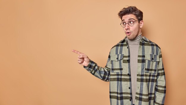 La foto de un chico sorprendido y sorprendido señala en el espacio de copia en blanco muestra el logotipo u oferta increíble usa gafas y camisa a cuadros aislados en beige con espacio de copia en blanco