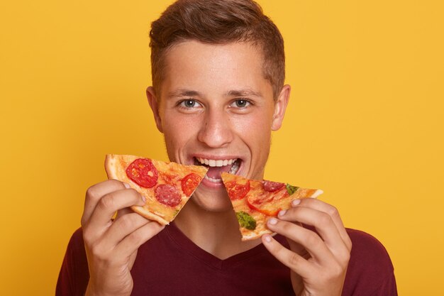 Foto de un chico vestido con una camiseta color burdeos que sostiene dos trozos de pizza y come comida rápida