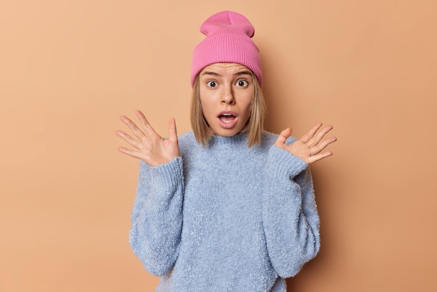 La foto de cintura arriba de una joven sorprendida mantiene la boca abierta con las palmas levantadas no puede creer que sus ojos se sientan estupefactos, usa un sombrero rosa y un suéter azul casual aislado sobre un fondo marrón Concepto de reacción