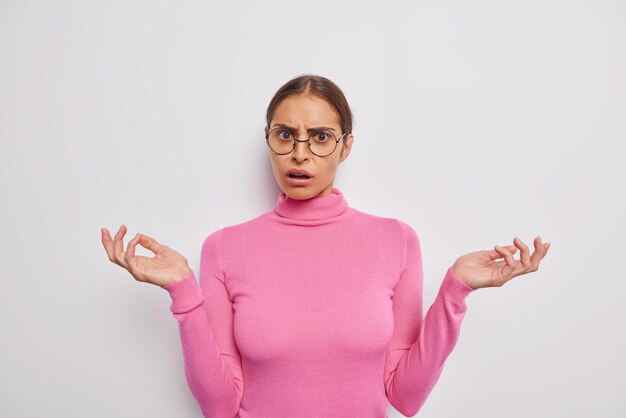 La foto de la cintura para arriba de una mujer bonita morena extiende las palmas tiene una expresión indecisa desconcertada que reacciona ante algo desagradable aislado sobre una pared blanca