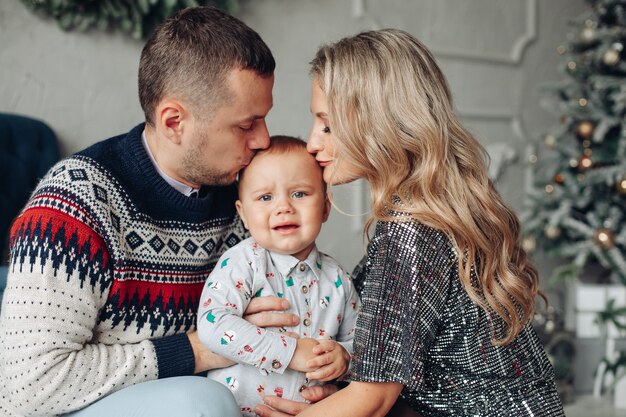 Foto de cintura para arriba de padres amorosos besando a su bebé en la cabeza con un árbol de Navidad