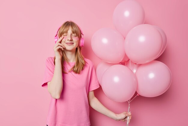 La foto de la cintura de una chica pelirroja aplica parches de belleza para el tratamiento de la piel mantiene los ojos cerrados escucha música a través de auriculares sostiene un montón de globos disfruta de la celebración de cumpleaños aislada en la pared rosa