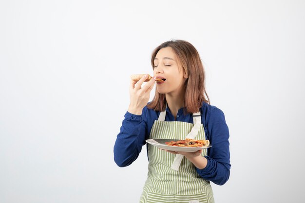 Foto de cocinera en delantal comiendo una rebanada de pizza en blanco