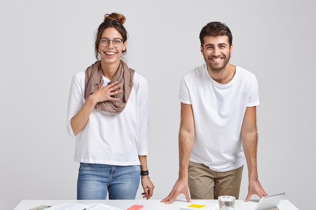 Foto de colegas felices que se regocijan en elogios del jefe por su buen trabajo, tienen sonrisas con dientes, se paran cerca del escritorio con tableta, vaso de agua y libros, aislados sobre una pared blanca