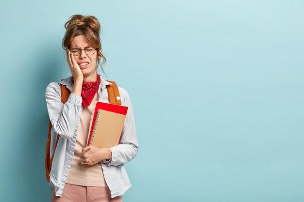 Foto de colegiala cansada estresante insatisfecha lleva el cuaderno y el libro en espiral, lleva gafas redondas