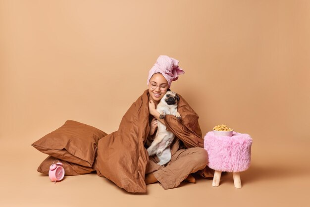 Una foto completa de una joven complacida abraza las poses de perro favoritas debajo de una manta suave se sienta en una pose de loto y tiene una relación amistosa cerca de almohadas, reloj despertador y una mesa pequeña con copos de maíz