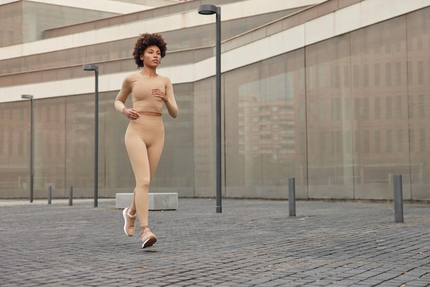 Una foto completa de una joven delgada y activa vestida con ropa deportiva y zapatillas deportivas corre rápido al aire libre tiene un entrenamiento matutino regular enfocado en la distancia La corredora atlética se prepara para el maratón