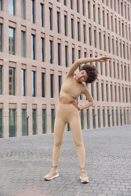 Una foto completa de una mujer activa de cabello rizado y delgado vestida con chándal se inclina hacia ejercicios al aire libre tiene poses de expresión segura de sí misma cerca de un edificio moderno tiene entrenamiento regular durante la mañana