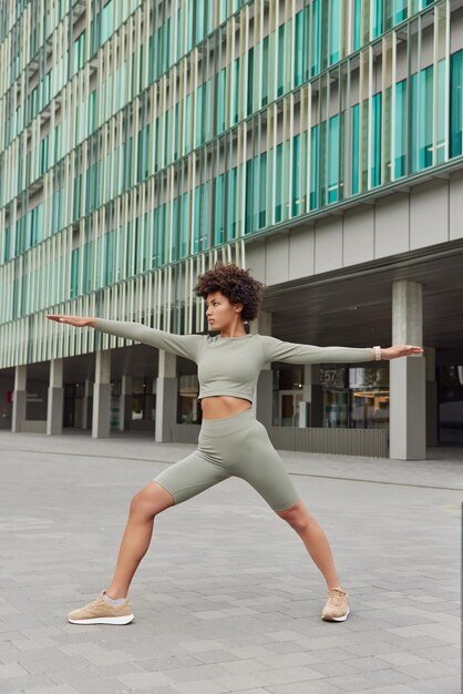 Una foto completa de una mujer deportiva motivada de pelo rizado estira ejercicios de brazos al aire libre vestida con poses de ropa activa cerca de un edificio urbano moderno enfocado en la distancia Concepto de entrenamiento y deporte