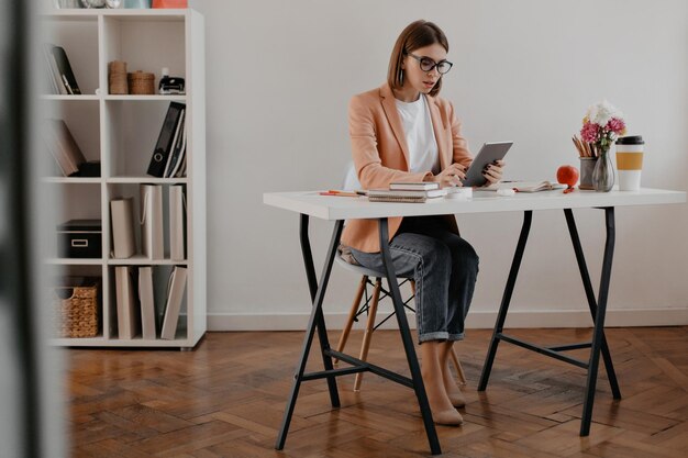 Foto completa de mujer de negocios con chaqueta rosa y jeans trabajando con iPad en su oficina