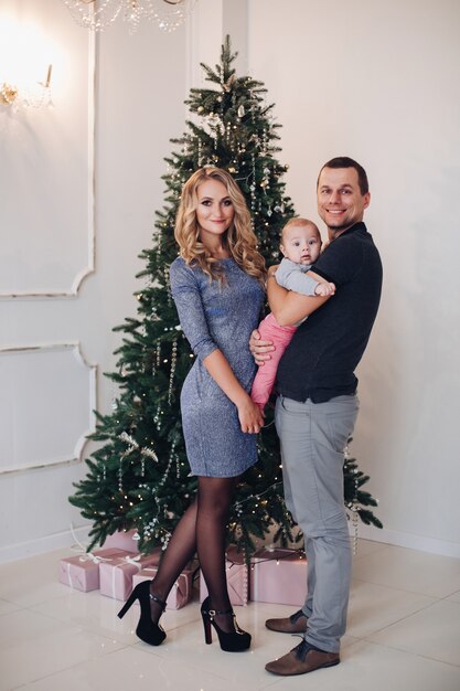 Foto de concepto de año nuevo de una familia joven feliz de dos padres y un bebé posando cerca del árbol de Navidad