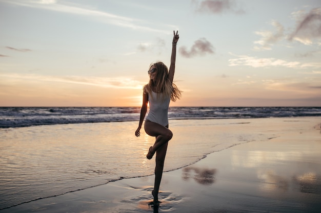 Foto de cuerpo entero de adorable chica delgada de pie sobre una pierna en la costa del océano.