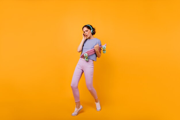 Foto de cuerpo entero de bailarina glamorosa con longboard. Retrato de mujer caucásica increíble posando en auriculares y escuchando música.