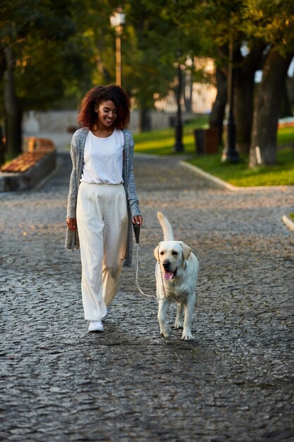 Foto de cuerpo entero de bastante saludable jovencita caminando por la mañana en el parque con perro