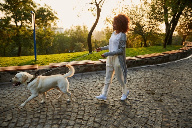 Foto de cuerpo entero de bastante saludable jovencita caminando por la mañana en el parque con perro