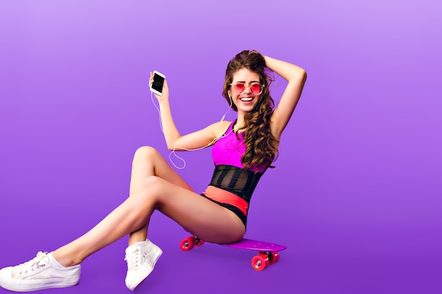 Foto de cuerpo entero de una chica atractiva con el pelo largo y rizado en gafas de sol rosa sentado en patineta sobre fondo púrpura. Viste traje de baño y escucha música con auriculares.