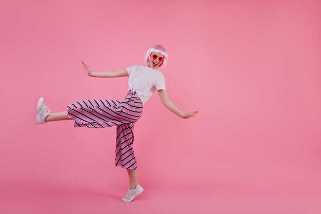 Foto de cuerpo entero de una chica con estilo increíble en pantalones rosas bailando. Retrato de mujer joven alegre en elegante periwig expresando emootions positivas