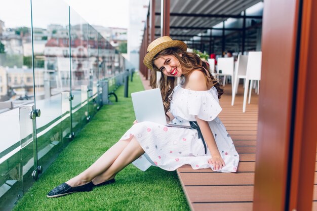 Foto de cuerpo entero de una chica guapa con el pelo largo sentada en el suelo de la terraza. Ella tiene una computadora portátil de rodillas y posando para la cámara