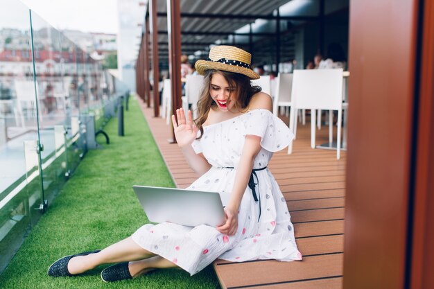 Foto de cuerpo entero de una chica guapa con el pelo largo sentada en el suelo de la terraza. Lleva un vestido blanco con hombros descubiertos, lápiz labial rojo y sombrero. Ella está hablando en una computadora portátil.