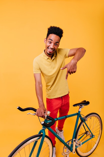 Foto de cuerpo entero de un chico negro sonriente en pantalones rojos divirtiéndose. Retrato de interior de hombre africano activo con bicicleta.