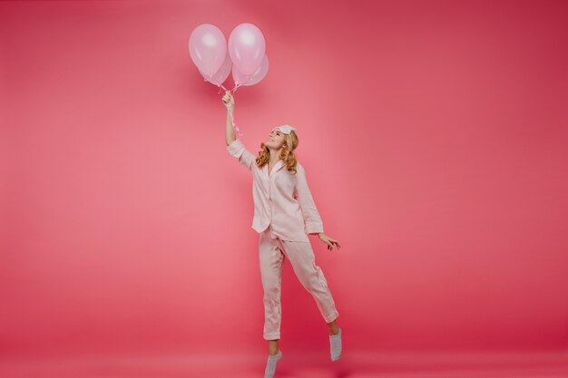 Foto de cuerpo entero de una entusiasta chica rubia posando con globos. Retrato de interior de dama alegre en máscara de dormir de puntillas en su cumpleaños.