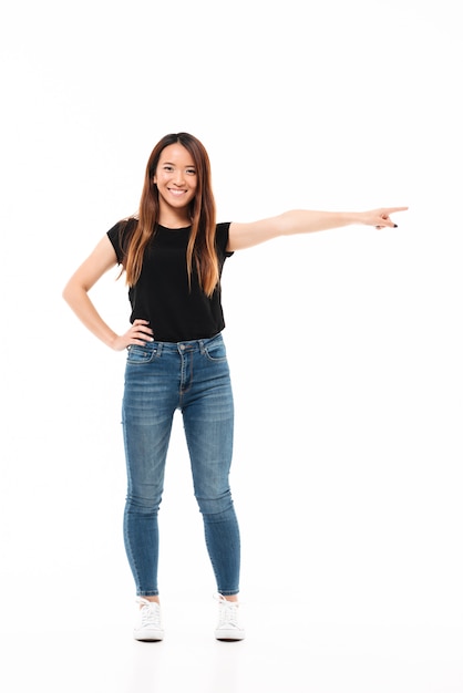 Foto de cuerpo entero de feliz mujer asiática encantadora en ropa casual de pie con la mano extendida, apuntando con el dedo, mirando a la cámara