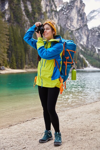 Foto de cuerpo entero de fotógrafo positivo toma una fotografía del río de montaña turquesa, posa en un hermoso lugar para la visita turística