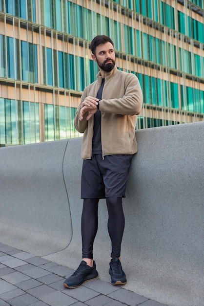 Foto de cuerpo entero de hombre barbudo pensativo en ropa deportiva espera a que el entrenador comience a trabajar, verifica el tiempo en el reloj de pulsera concentrado frente al entorno urbano. Concepto de personas y estilo de vida