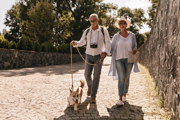 Foto de cuerpo entero de hombre con camisa blanca y jeans con cámara y mujer con sombrero y blusa azul a rayas con tarjeta y corgi caminando en el parque.