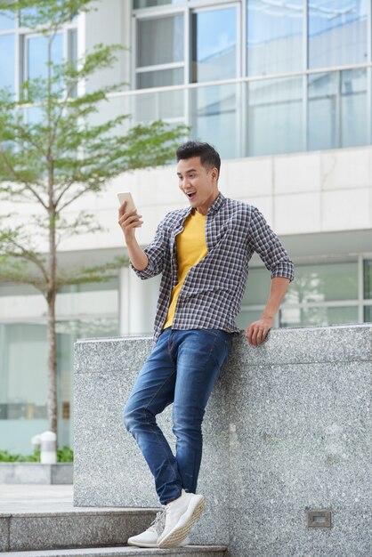 Foto de cuerpo entero del joven apuesto excitado por la noticia leída en el teléfono inteligente