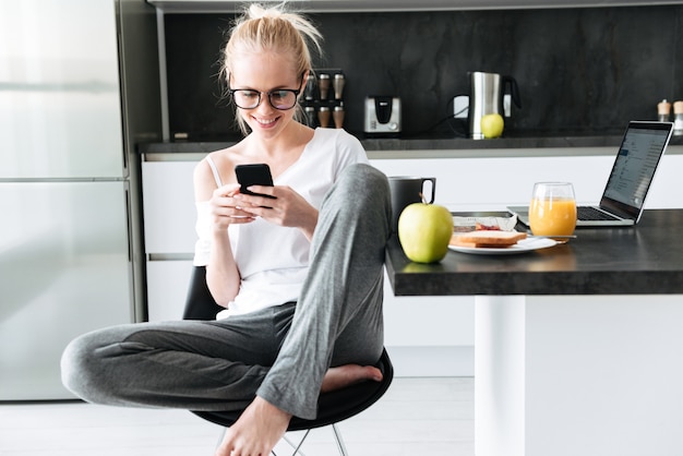 Foto de cuerpo entero de joven enfocada usando un teléfono inteligente mientras está sentado en la cocina