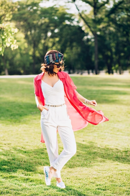 Foto de cuerpo entero de una linda chica morena en pañuelo de pie en el parque de verano. Viste ropa blanca, gafas de sol y camisa larga rosa. Mantiene la mano en el bolsillo y mira hacia abajo.