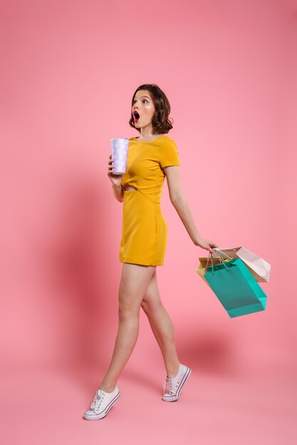 Foto de cuerpo entero de linda mujer sorprendida en vestido amarillo con bebida y coloridos bolsos de compras, mirando a un lado