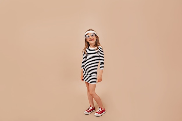 Foto de cuerpo entero de linda niña encantadora con vestido, gorra y zapatillas de deporte posando en la cámara sobre fondo beige