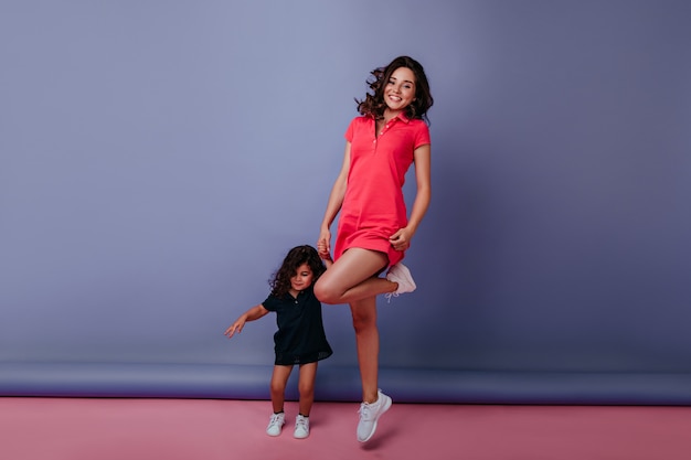 Foto de cuerpo entero de una magnífica mujer joven bailando con su hija y sonriendo. Señora bien vestida cogidos de la mano con la niña en la pared púrpura.