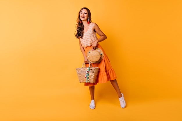 Foto de cuerpo entero de mujer inspirada con accesorios de verano. Modelo femenino de jengibre feliz en falda naranja con sombrero y bolso.