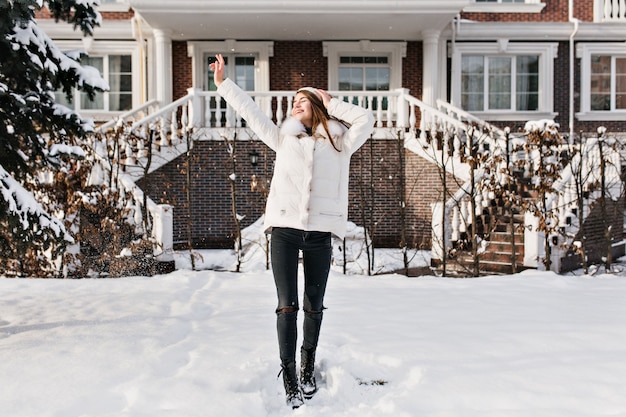 Foto de cuerpo entero de una mujer joven delgada en ropa de abrigo con estilo disfrutando del fin de semana de invierno. Retrato al aire libre de fascinante dama caucásica en pantalones oscuros posando con las manos en alto en un día frío.