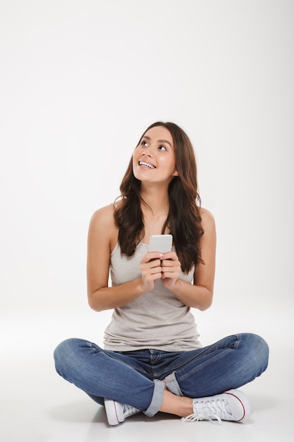 Foto de cuerpo entero de una mujer joven en jeans y zapatillas de deporte sentado con las piernas cruzadas en el piso con un teléfono inteligente plateado en la mano, sobre una pared blanca